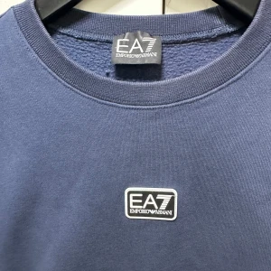 Blå sweatshirt från EA7 Emporio Armani - Snygg blå sweatshirt från EA7 Emporio Armani med rund hals och ett stilrent logomärke på bröstet. Tröjan har lång ärm och är tillverkad i mjuk bomullsmix som känns skön mot huden. Perfekt för dig som gillar enkel och clean streetstyle.