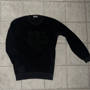 Svart Kenzo tröja med tigerbroderi - Svart tröja från Kenzo i storlek M med ikoniskt tigerbroderi framtill och Kenzo Paris-text. Rund halsringning och ribbade muddar vid ärmslut och nederkant. Perfekt för dig som gillar stilrena designer med en twist.passar också för storlek S !