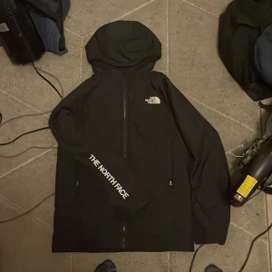 Svart vindjacka från The North Face - Svart vindjacka från The North Face i storlek XL i barnstorlek(14-16yr) med huva och dragkedja framtill. Jackan har logotypen tryckt på bröstet och längs ena ärmen. Perfekt för dig som vill ha en clean och sportig look med street-vibe.