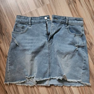 Blå jeanskjol med slitningar - Snygg NY  blå jeanskjol , strl XL , med rå kant och slitna detaljer framtill och på bakfickorna. Kjolen har klassiska fickor, bälteshällor och dragkedja framtill. Stretchig .