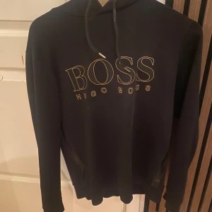 Svart hoodie från Hugo Boss - Svart hoodie från Hugo Boss med stor guldfärgad logga på bröstet. Klassisk huva med snörning och långa ärmar. Perfekt för dig som gillar stilrena och trendiga plagg med premiumkänsla.