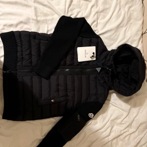 Svart Moncler dunjacka med huva - Svart dunjacka från Moncler med ribbstickade ärmar och huva. Jackan har quiltad kropp, dragkedja framtill och en ficka med knapp. Moncler-logga på ärmen och snygga detaljer. Perfekt för dig som gillar stilren och sportig look.