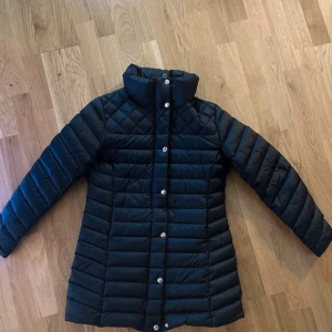 Svart dunjacka från GANT, M - Snygg svart dunjacka från GANT i storlek M. Jackan har quiltad design, hög krage och silverfärgade knappar framtill. Långärmad och med GANT-logga broderad på ryggen. Perfekt för kalla dagar med stilren look.