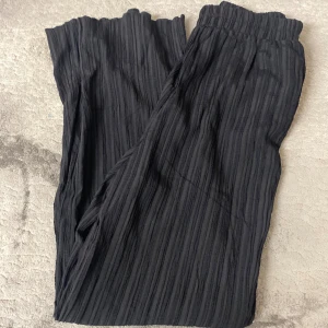 Svarta plisserade byxor med resår - Svarta byxor med plisserad struktur och resår i midjan. Vida ben och mjukt, lätt material som ger en avslappnad känsla. Perfekta att styla med en enkel topp eller hoodie.