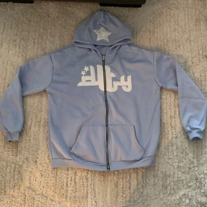 Ljusblå hoodie med stjärnor och tryck - Ljusblå hoodie från Ducty med vit broderad stjärna på huvan och stort vitt tryck med stjärnor på bröstet. Tröjan har dragkedja framtill, två fickor och ribbade muddar. Perfekt för dig som gillar streetwear och snygga detaljer.