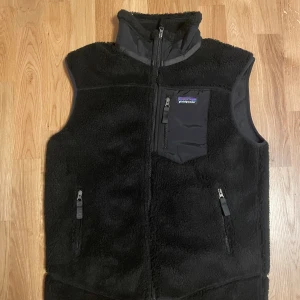 Svart fleeceväst från Patagonia - Svart fleeceväst från Patagonia med hög krage och tre fickor med dragkedja, varav en bröstficka med logotyp. Västen har en mjuk och fluffig yta, meshfoder på insidan och detaljer i syntetmaterial. Perfekt för lager-på-lager och outdoor.