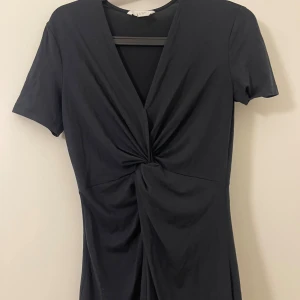 Svart kort omlottklänning med twist - Svart kort klänning från H&M med snygg twistdetalj framtill och djup v-ringning. Klänningen har korta ärmar och är gjord i mjuk bomull, vilket gör den både bekväm och stilren. Perfekt för dig som gillar minimalistisk och trendig look.
