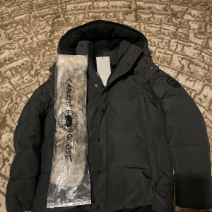 Canada Goose - Svart dunjacka från Canada Goose med huva och avtagbar pälsdetalj. Jackan har diskret logga på ärmen, dragkedja och knappar framtill samt flera fickor. Tillverkad i slitstark polyester och fodrad med dun och fjädrar för extra värme. Perfekt för kalla vinterdagar. (finns i också i stl L)