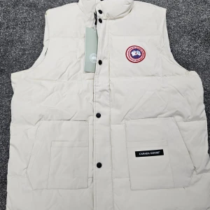Beige dunväst från Canada Goose - Snygg beige dunväst från Canada Goose med klassisk logga på bröstet. Västen har två stora fickor framtill, svarta knappar och hög krage. Insidan är svart och materialet känns slitstarkt och varmt. Perfekt för lager-på-lager under kyliga dagar.