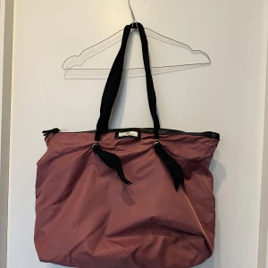 Rosa shoppingväska från DAY Birger et Mikkelsen - Stor och rymlig shoppingväska i mörkrosa polyester med svarta handtag och detaljer. Väskan har dragkedja upptill och coola knutdetaljer på handtagen. Perfekt för dig som vill ha plats för allt du behöver under dagen.