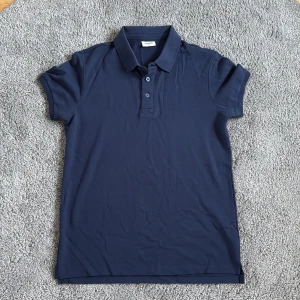 Marinblå pikétröja från Kappahl - Snygg marinblå pikétröja från Kappahl med klassisk krage och tre knappar framtill. Tröjan är kortärmad och tillverkad i mjuk bomull, perfekt för en stilren och avslappnad look.