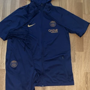 Marinblå PSG kofta från Nike med byxor - Snygg marinblå kofta och byxor från Nike x PSG med guldfärgade detaljer och loggor. Jackan har huva, hel dragkedja och sidofickor med dragkedja. Perfekt för dig som gillar fotboll och vill ha en sportig stil.