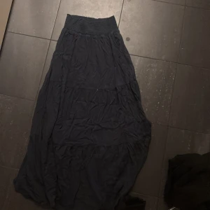 Mörkblå långkjol från 157 XS - Mörkblå långkjol från 157 i storlek XS. Kjolen har en bred, smockad midja som sitter skönt och ger en snygg siluett. Materialet är mjukt och lätt, perfekt för dig som gillar enkel och stilren look. Passar dig som vill ha en flowy och bekväm kjol med lite volym.