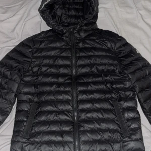 Svart pufferjacka Moose Knuckles  - Svart pufferjacka från Moose Knuckles i storlek S/xs Jackan har huva, två sidofickor med dragkedja och ett snyggt logomärke på ärmen. Quiltad design och hel dragkedja framtill. Perfekt för kalla dagar och har en modern, clean look. Den passar mig som brukar ha S