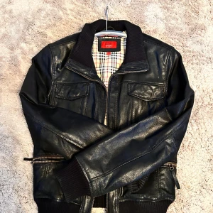 Svart skinnjacka från Kaos by Auluna - Snygg svart skinnjacka från Kaos by Auluna med rutigt innerfoder. Jackan har ribbade muddar, dragkedja framtill, två bröstfickor med lock och coola detaljer vid ärmslut och midja. Perfekt för dig som vill ha en klassisk men edgy look.