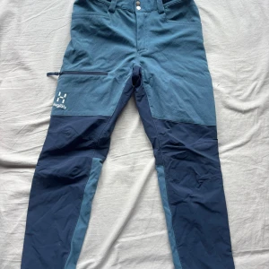 Blå täckbyxor från Haglöfs Junior 140 - Säljer ett par blå täckbyxor från Haglöfs i storlek Junior 140. Byxorna har två olika blå nyanser, förstärkta partier och praktisk dragkedja på benet. Materialet är slitstarkt och passar perfekt för utomhusaktiviteter. Snygg Haglöfs-logga på benet.