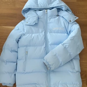 Ljusblå pufferjacka från WAWA - Säljer en riktigt snygg ljusblå pufferjacka från WAWA med broderad logga på bröstet och ryggen. Jackan har huva, dragkedja framtill och två fickor med dragkedja. Tillverkad i polyester med bomullsfyllning, perfekt för kalla dagar.