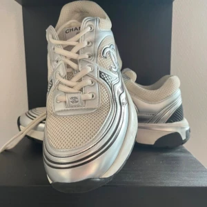 Chanel sneakers i silver och vitt - Snygga Chanel sneakers i silver och vitt med mesh-överdel och metallic-detaljer. Ikonisk CC-logga på sidan och sulan, samt svarta och vita inslag på yttersulan. Skorna har snörning och en rund tå, perfekt för dig som gillar lyxiga och trendiga sneakers.