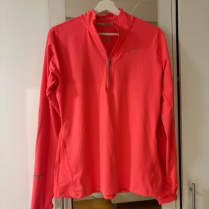 Korallfärgad långärmad träningströja Nike - Snygg neonrosa långärmad träningströja från Nike med halv dragkedja framtill och diskret logga på bröstet. Tillverkad i ett lätt och stretchigt funktionsmaterial som andas. Perfekt för löpning eller gym. Reflekterande detaljer på ärmen.
