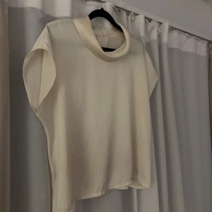 Krämvit blus med korta ärmar - Säljer en stilren krämvit blus med korta ärmar och hög krage. Blusen har en mjuk, slät yta och stängs med dragkedja i nacken. Passformen är normal och tyget känns lätt och luftigt, perfekt för ljusa dagar.