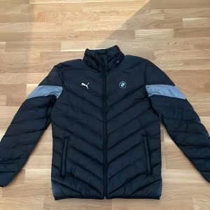 Svart BMW Motorsport pufferjacka Puma - Svart pufferjacka från Puma i samarbete med BMW Motorsport. Jackan har gråa detaljer på ärmarna, broderade loggor fram och bak, samt dragkedja framtill. Materialet är syntet och jackan har en modern, sportig look med hög krage och två sidofickor.