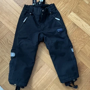 Svarta täckbyxor Polarn O. Pyret 104 - Svarta täckbyxor från Polarn O. Pyret med justerbara hängslen, reflexdetaljer och dragkedjor vid fickorna. Insidan är fodrad med mjuk fleece . Stl 104