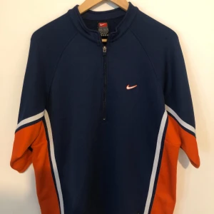 Nike half zip tröja marinblå & orange - Sportig marinblå tröja från Nike med orange och vita detaljer längs ärmarna. Tröjan har half zip framtill och korta ärmar, perfekt för träning eller chill. Snygg logga på bröstet och cool retrostil.