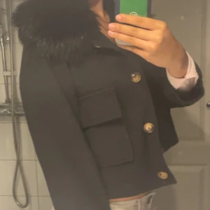 Svart kappa med fuskpäls och guldknappar - Snygg svart kappa med stor krage i fuskpäls och guldiga knappar framtill. Jackan är croppad och har två stora fickor med lock. Insidan har ett coolt mönstrat foder. Perfekt för dig som vill sticka ut med en klassisk men trendig look. Vet inte riktigt vilken storlek, typ en S eller XS