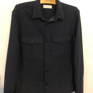 Mörkgrå overshirt från Jack & Jones - Snygg mörkgrå overshirt från Jack & Jones Premium med klassisk krage och två bröstfickor med knappar. Skjortan har långa ärmar och är tillverkad i ett mjukt material som ger en stilren look. Perfekt att bära som lager över t-shirt eller hoodie.