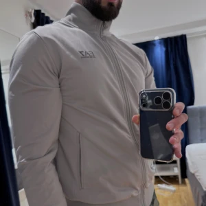 Grå EA7 track jacket med dragkedja - Snygg grå track jacket från EA7 Emporio Armani med hög krage och hel dragkedja framtill. Jackan har två fickor med dragkedja och diskret logga på bröstet. Tillverkad i mjuk polyester för en sportig och clean look. Storlek XL men skulle säga att den även passar om du är L