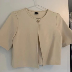 Beige kort cardigan  från Gina tricot  - Stilren beige cardigan  från Gina tricot med kort ärm och rund halsringning. Koftan  är kort och stängs med en dekorativ knapp framtill. Perfekt för lager-på-lager och enkel att matcha med olika outfits.
