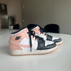 Nike Jordan 1 Mid rosa/svart/vit - Nike Jordan 1 Mid sneakers i en snygg färgkombo av ljusrosa, svart och vit. Skorna har klassisk snörning, rund tå och platt sula i gummi. Ikonisk Jordan-logga på sidan och Nike Swoosh i rosa. Ovandel i skinn med perforerad tå för extra ventilation.