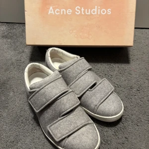  Acne Studios sneaker - Snygga grå sneakers från Acne Studios med mjuk mocka-look och breda kardborreband. Skorna har rund tå, vit platt sula och diskret Acne Studios-logga på sidan. Insidan är vit och ger en clean kontrast mot det grå yttre. Perfekt för dig som gillar minimalistisk stil.