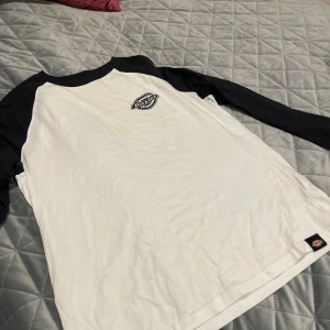 Svartvit långärmad tröja Dickies XS - Snygg svartvit långärmad tröja från Dickies i storlek XS. Tröjan har svart raglanärmar och vit kropp med Dickies-logga tryckt på bröstet. Rund halsringning med svart kant. Perfekt för dig som gillar streetwear och enkel stil. Små fläckar på framsidan nedtill.