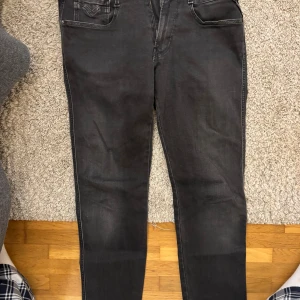 Replay svarta jeans Anbass tapered - Svarta Replay Anbass jeans med tapered fit och klassisk femficksdesign. Snygga detaljer på bakfickan och läderpatch i midjan. Jeansen är tillverkade i stretchigt bomullsmaterial för extra komfort och har dragkedja i gylfen.
