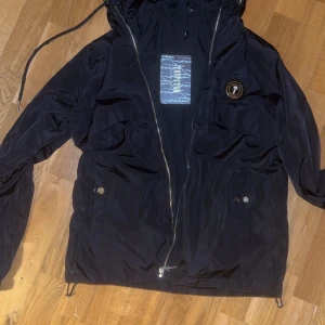 Trapstar windbreaker - Svart vindjacka från Trapstar med huva, dragkedja och snörning i nederkant. Jackan har broderad Trapstar-logga på ryggen och en rund patch på bröstet. Tillverkad i lätt polyester, perfekt för blåsiga dagar. Snygg streetstil med praktiska fickor och coola detaljer. Pris går att diskutera