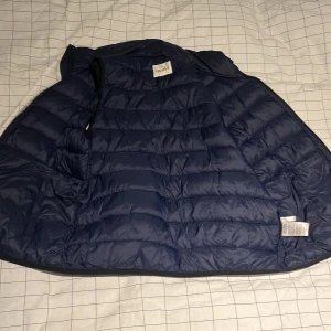 Mörkblå dunväst från KappAhl - Snygg mörkblå dunväst från KappAhl i storlek 164. Västen har quiltad design, två fickor med dragkedja och hög krage. Perfekt att slänga över en hoodie när det är kyligt ute. Materialet är polyester och dun, vilket gör den både lätt och varm.