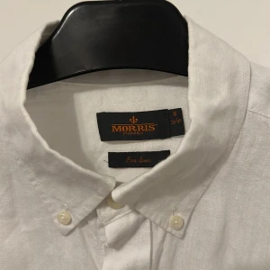 Vit linneskjorta från Morris M - Fräsch vit skjorta från Morris i ren linne, med klassisk button-down krage och snygga knappar framtill. Liten svart broderad logga på bröstet och diskret Morris-märke vid manschetten. Perfekt för dig som gillar stilrena och luftiga plagg.