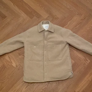 Beige teddyjacka från Weekday, S - Beige teddyjacka från Weekday i storlek S. Jackan har två stora fickor framtill, klassisk krage och stängs med knappar. Insidan är fodrad med mjuk vit teddy, perfekt för kyliga dagar. Enkel och stilren design som passar till det mesta.