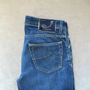 Blå jeans från Jacob Cohen - Snygga blå jeans från Jacob Cohen med klassisk femficksdesign och kontrastsömmar. Jeansen har en rak passform och är tillverkade i slitstarkt denim med en mörk tvätt och lätt slitning på benen. Bakfickan har en unik form och logopatch i mocka.