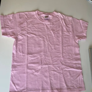 Ljusrosa t-shirt från Fruit of the Loom - En basic ljusrosa t-shirt från Fruit of the Loom i 100% bomull. Klassisk rund halsringning och korta ärmar. Perfekt enkel stil och skön passform för dig som gillar en clean look.