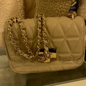Beige quiltad axelväska med guldkedja - Snygg beige axelväska med quiltat mönster och guldfärgad kedja som axelrem. Väskan har en rektangulär form och ett stilrent lås i guld på framsidan. Materialet är skinnimitation och insidan har ett praktiskt fack för småsaker.