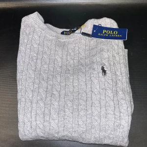 Grå kabelstickad tröja  - Snygg grå kabelstickad tröja med klassisk rund halsringning och broderad logga på bröstet. Perfekt för lager-på-lager och passar till allt. Mjuk och skön känsla i stickad bomull.