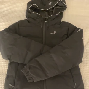 Svart vinterjacka från Pelle P - Svart vinterjacka från Pelle P med huva, dragkedja och snygga reflexdetaljer. Jackan har logga på bröstet och ärmen, samt praktiska fickor med dragkedja. Perfekt för kalla dagar när du vill hålla stilen och värmen.