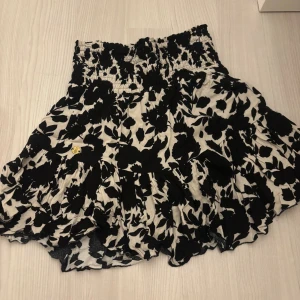 Blommig kortkjol från Pernilla Wahlgren - Supersöt kortkjol från Pernilla Wahlgren med svartvitt blommönster och volangkant. Kjolen har smock i midjan för en bekväm passform och är perfekt för dig som gillar en feminin och trendig stil. Lätt och luftig känsla, passar perfekt till sneakers eller sandaler. Pris kan diskuteras!