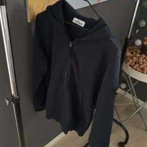 Mörkblå hoodie från Stone Island Junior - Svart hoodie från Stone Island Junior med klassisk logotyp-patch på ärmen. Tröjan har huva, dragkedja framtill och två fickor. Materialet är mjuk bomull och passformen är normal. Perfekt för dig som gillar streetwear och snygga detaljer. Köpt på flannels i london. Har även kvitto. Passar dig som är mellan 170-180
