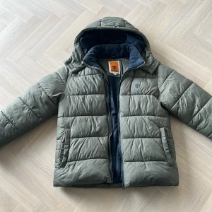 Grön pufferjacka från Timberland - Säljer en grön pufferjacka från Timberland med huva och dragkedja. Jackan har quiltad design, två fickor framtill och diskret Timberland-logga på bröstet. Perfekt för kyliga dagar och riktigt skön att bära. Fodrad insida i mörkblått tyg. Säljs pågrund av att den är för liten, pris kan diskuteras 