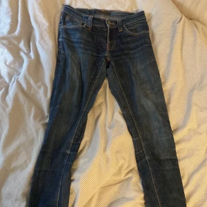 Blå skinny jeans från Nudie Jeans - Säljer ett par klassiska blå skinny jeans från Nudie Jeans med snygga orange sömmar och ikonisk bakficka. Jeansen har normal passform, fem fickor och är tillverkade i mjukt denimtyg. Perfekta för dig som gillar en smal siluett och stilren look.
