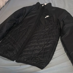 Svart quiltad pufferjacka från Nike - Nike pufferjacka i svart med quiltad design och dragkedja framtill. Jackan har långärmade ärmar, hög krage och vit Nike-logga på bröstet. Materialet är syntet och jackan har Therm-Fit för extra värme. Perfekt för kalla dagar.