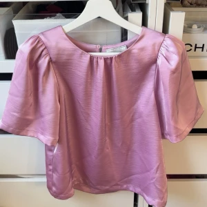 Rosa blus i satin med puffärm från Neo Noir - Supersöt rosa blus i glansigt satinmaterial med korta puffärmar och rund halsringning. Blusen har en avslappnad passform och knäppning bak i nacken. Perfekt för dig som vill ha en feminin och trendig look. 